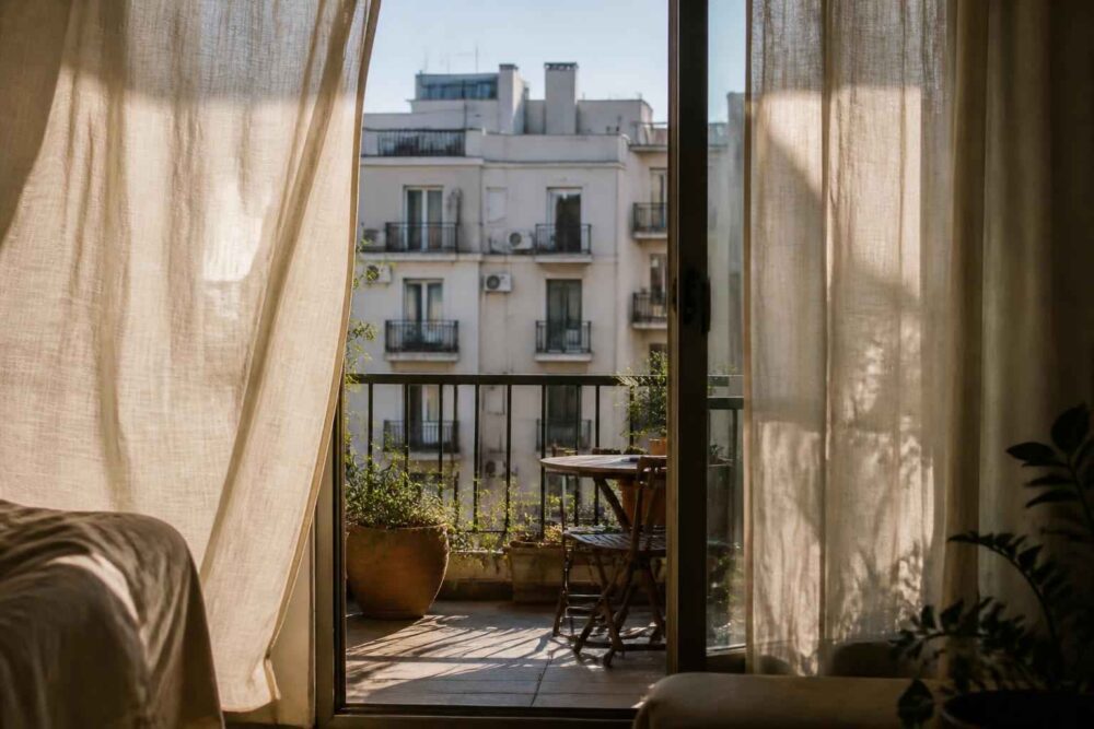 Słoneczny balkon z roślinami i stolikiem, widoczny przez otwarte drzwi z wnętrza mieszkania